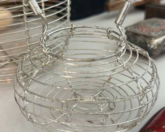 Silver Wire Basket Small Handle — Small decorative silver-tone wire basket with a wrapped wire handle and open, coiled construction. Good display piece for trinkets, potpourri, or as tabletop decor; lightweight and shows minor surface wear consistent with use. No visible maker's marks; handle is firmly attached and the coil wire construction is intact. Measures compactly and is easy to move or incorporate into grouped displays.