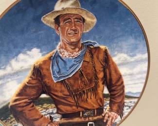 John wayne plate — Decorative round collector plate featuring a painted western cowboy in hat and fringe jacket with a blue neckerchief; metal rim and hanging clips are visible. The plate is a wall-hang style decorative piece suitable for western-themed decor or collections. From photos the painted surface and rim show no obvious chips or cracks but there is light surface wear consistent with display. Measures approximately 10–12 inches in diameter (see dimensions) and sold as a single decorative plate.