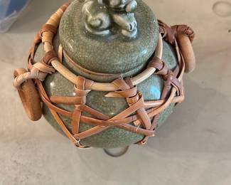 celadon-glazed lidded jar — Celadon-glazed ceramic lidded jar with a small monkey or cherub-style finial and a woven bamboo carrier. The crackle-glaze surface and muted green tone are visible across the body and lid; bamboo wrapping with wooden buttons secures the piece. Underside shows a hand-applied mark (photo slightly blurred) and a small price sticker; no obvious chips or cracks visible in provided photos. Compact decorative jar perfect for shelf display or as a small storage container.