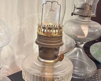 Glass Kerosene Oil Lamp — Clear pressed-glass kerosene oil lamp with tall glass chimney and metal burner assembly. The lamp has a faceted round font and a crown-style brass burner with adjustable wick knob; chimney shows light soot residue and burner has surface patina consistent with age. Good decorative condition for use or display after cleaning; no obvious chips or cracks visible in the glass from photos. Measures compactly and is a classic functional accent for a mantel or table.