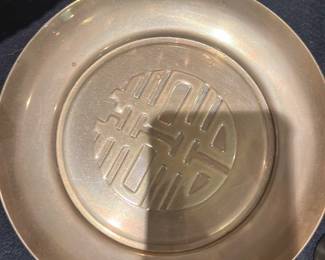Brass DecorativeDish — Heavy brass decorative ashtray or small catchall dish with a raised circular center featuring an Asian-inspired longevity/shou-style motif. The piece shows a warm patina and light surface scratches consistent with age and use but no major dents or repairs visible. Solid, usable accent for a coffee table or shelf; measures small and is substantial in hand. Good vintage condition with even wear to the finish.