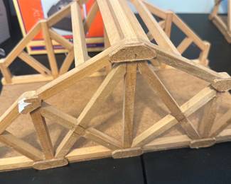 Wooden Truss Bridge Model — Handcrafted wooden truss bridge model constructed from small timber strips glued into a triangular truss pattern on a rectangular base. The piece appears to be a scale or educational model showing basic roof/bridge truss geometry; unfinished wood with visible glue joints and light wear at several corners. Sturdy for display or classroom demonstration but shows cosmetic imperfections consistent with handling. Measures roughly 14 inches long and low-profile for tabletop display.