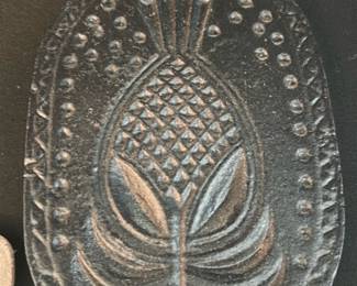 Cast Iron Pineapple Trivet — Heavy cast-iron trivet featuring a raised pineapple motif and decorative border; a functional vintage-style piece for protecting surfaces from hot pans. Solid construction shows light surface wear and mild surface rust/patina consistent with age and use; small drilled hole at top for hanging. Measures compactly for countertop use and works well as kitchen decor or a practical trivet.