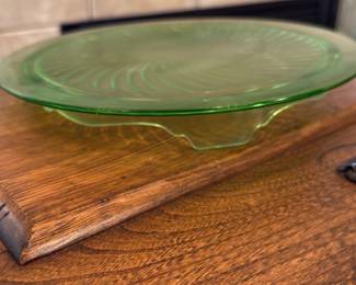 Green Depression Glass Cake Plate — A green Depression-era style glass cake plate with a molded swirl center and three scalloped feet. The piece displays the characteristic translucent green (uranium-style) color and pressed-glass patterning seen in 1930s–1950s production; it's solidly made with light surface wear visible in close-up photos but no chips or cracks apparent. Use as a dessert stand or display piece; measures approximately 10–11 inches across and is lightweight enough for everyday use. Good condition overall with minor surface scratches consistent with age and use.