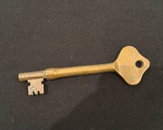 Brass Skeleton Key Large — Solid brass skeleton key shown from multiple angles; long single-bit design typical of old warded locks or decorative reproductions. Heavy-feeling solid metal construction with patina and light surface wear consistent with age or use; no visible maker name beyond small stamped mark on bow (not legible in photos). Useful as a hardware replacement, decorative piece, or key for vintage cabinets/doors; pictured on a living-room surface for scale.