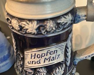 Hopfen und Malz German Lidded Stein — Decorative German-style stoneware beer stein labeled "Hopfen und Malz" with relief hop and barley decoration and a pewter hinged lid. The cobalt-blue glaze contrasts with beige relief details; handle and thumb lift are intact and the lid shows light surface wear consistent with age. A classic decorative stein suitable for display or collectors of beerware; no visible chips or cracks in photos but sold as used. Measures approximately 8 inches tall overall (including lid).