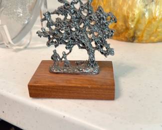metal tree figurine on wood base — Small sculptural tree figurine cast in a silvery metal and mounted on a rectangular walnut-finish wood base. The textured, branchy tree silhouette includes two tiny human figures at the trunk and shows a hand-cast, decorative look suitable for a shelf or desk. Base underside has felt padding; overall condition is good with minor surface wear consistent with display. Measures small and lightweight—an easy accent piece for a living room or office.