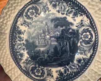 Blue Transferware Decorated serving Plat — Blue transferware decorative plate with a central pastoral/architectural scenic transfer and ornate floral border, set in a textured basketweave rim. Underside shows a blue printed crest/coat-of-arms mark (no readable maker name visible in photos). Plate appears to be ceramic/earthenware with crazing visible in glaze and light surface wear consistent with age. A good decorative or display piece for dining-room or curio cabinet; measurements and weight are estimated from photos.