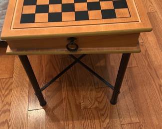 Wood Top Chess & Backgammon Table — This multifunction game table features a wood top with a chessboard inlay and backgammon pattern on the underside of the sliding lid, perfect for games in the master bedroom. The top lifts to reveal an internal storage compartment lined in black felt containing backgammon/checkers pieces and accessories stored in plastic bags. Table stands on black metal legs with stretcher supports and is a compact, lightweight occasional table suitable for small spaces. It is in good condition with minor surface wear and light scuffs from normal use; all game pieces appear present though unboxed.