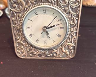 Ornate Table Clock — Small decorative metal tabletop clock with an ornate raised floral scroll border and a round white dial with Roman numerals. The face is marked "QUARTZ" and the clock shows light surface tarnish and minor wear consistent with age but appears intact; battery-powered movement (battery not tested). Compact decorative piece suitable for mantel, desk, or bedside table.