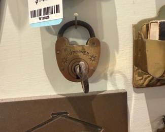 Improved Brass Padlock Wall Hook — Small brass padlock-style wall hook stamped "IMPROVED" with decorative star motifs and an integrated iron shackle and hook. The piece appears to be a novelty or decorative repurposed lock used as a hanging hook; finish shows patina and light surface wear consistent with age. Solid metal construction and compact size make it useful for keys or light items; mounting hardware not visible but a nail is shown holding it in place. Good condition cosmetically with expected oxidation on the shackle and hook.