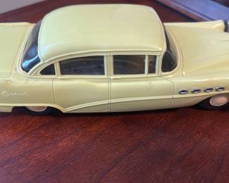 YellowBuick Toy Car — Die-cast style yellow metal toy car modeled after a classic Buick, offered from an office room estate sale. The body is painted yellow with chrome-colored bumpers and grille, clear plastic windows, and a stamped metal chassis that shows surface rust and patina on the underside. Approximately 9 inches long (see dimensions) and lightweight, this collectible toy shows wear consistent with age including scuffs, paint chips, and oxidation on the chassis but intact wheels and body. Good display piece for a classic car or toy collection; sold as-is with visible cosmetic wear and vintage character.