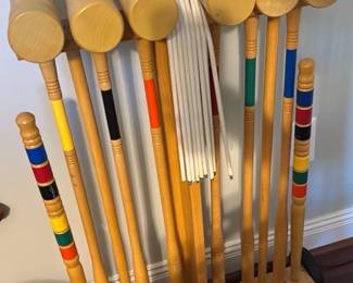Wooden Croquet Set with Stand — Classic full-size wooden croquet set complete with six painted balls, six mallets, six color-banded wickets, and a wooden carrying stand with wheels and handle. Brightly painted balls and color-coded mallet shafts match standard backyard croquet sets; stand stores mallets vertically and holds balls neatly at the base. Good used condition with surface scuffs and light wear to paint consistent with outdoor play; all parts appear present as shown. Compact wheeled stand makes it easy to move and store when not in use.