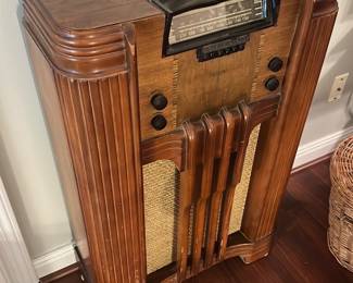Philco Art Deco Console Radio — Philco wood console radio in an art-deco style with stepped fluting and rounded corners, featuring the original tuning dial and push-button selectors. The cabinet shows attractive figured veneer and woven speaker grille cloth; hardware and knobs appear present though functionality is untested. A solid display piece for restoration or vintage audio enthusiasts—cabinet has some surface wear and light scuffs consistent with age. Measures roughly like a floor console and will need standard care if you plan to power or refinish it.