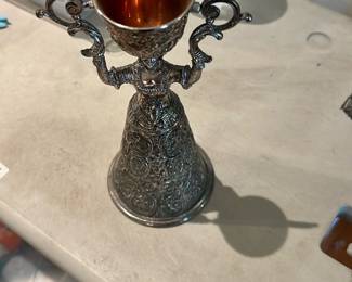 Nuremburg Bridal Wedding Cup — Ornate Two‑Handled Metal Chalice is a decorative metal cup with intricate all-over engraved or repoussé ornament and a flared bell-shaped base. The piece features two scrolled handles and a gold-toned interior liner visible in the photos; overall styling suggests a decorative or ceremonial chalice. Surface shows expected light tarnish and age-appropriate wear to the exterior finish but no obvious dents or breaks visible in the images. Compact and eye-catching, it works well as a mantel accent or collectible metalwork piece.
