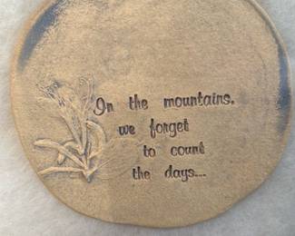 Ceramic Wall Plaque — Decorative round hanging plaque stamped with the phrase "On the mountains, we forget to count the days..." and a simple floral motif. Made to hang by a twine loop, the piece has a rustic, lightly distressed finish and appears intended for casual wall display or cabin decor. Condition is good with intentional surface wear as part of the finish and no visible cracks or major damage. Measures small and lightweight — an easy accent for a bedroom, hallway, or entry.