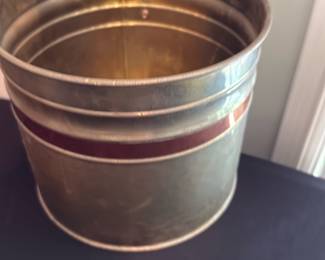Brass Container — Heavy brass decorative wastebasket or small planter with a simple ribbed band and a narrow red-brown inlay around the middle. Solid metal construction with visible surface wear, light dents, and patina consistent with age; interior shows spotting and oxidation. Good decorative condition for use as a planter, umbrella stand, or accent piece; measures roughly tabletop size and is sturdy but cosmetically worn.