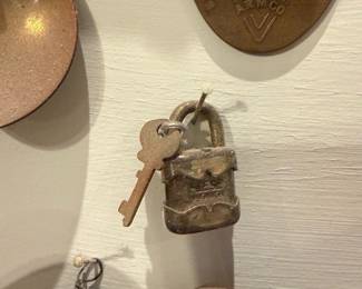 Brass Decorative Padlock with Key — Small brass padlock with its original single key shown hanging on a display board. The lock shows a scalloped decorative front and surface verdigris and wear consistent with age; maker name stamped faintly on the face but partially worn. Solid small hardware piece suitable for collection or decorative use; key is included. Condition is fair with corrosion and surface wear visible but intact structure.