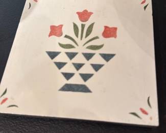 Floral Tile Trivet — Small square ceramic trivet with a hand-decorated floral motif in red, green and blue on a white ground and a black slate or stone backing with four felt-topped wood feet. Useful as a hot pad or decorative tabletop accent; shows light surface wear and slight edge wear visible in photos but overall sound. Compact, simple design that works in country, cottage, or farmhouse style kitchens. Backing keeps surfaces protected and feet appear intact.