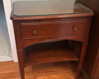 Mahogany Nightstand with Glass Top — Mahogany nightstand featuring a single drawer with round brass pulls and a lower display shelf, topped with a fitted glass protector. Solid-appearing hardwood construction with a warm reddish-brown finish and simple curved apron detailing that suits traditional or transitional bedrooms. Overall compact footprint makes it ideal as a bedside table or small end table; measurements listed below are estimates. Good estate-sale condition with light surface wear and minor scuffs consistent with normal use; glass top appears intact with no visible chips. Classic, well-made piece that offers practical storage and display in the master bedroom.