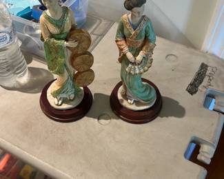 Asian Lady Porcelain Figurine — Pair of porcelain/ceramic Asian lady figurines on round wooden bases, each depicting a woman in traditional dress holding accessories (one with fans, one with a basket). Both show hand-painted detail in greens, golds and creams with molded hair and delicate facial features; bases are lacquered wood. Good overall condition with light shelf wear and small paint touchwear consistent with age and display visible in photos. Each stands roughly tabletop size and they make a decorative pair for a mantel or curio cabinet.