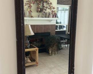 Ornate Carved Wall Mirror — Decorative carved wall mirror with ornate floral and ribbon motifs around the frame. The frame appears to be painted/gilded wood or composition with subtle polychrome accents in green, red and gold and a textured background finish. Measures approximately 36 inches tall by 14 inches wide and about 1.5 inches deep, a good size for an entryway or hallway. Mirror glass shows light age-related spotting and the frame has minor nicks and paint loss consistent with normal wear but is structurally sound.