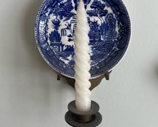 Blue Willow Wall Sconce — Decorative blue willow plate wall sconce with a single brass-finish candle arm, perfect for adding traditional charm to a living room. The piece features a blue and white transferware plate mounted on a metal bracket with a curved arm and round drip ring, holding a twisted taper candle. Overall wall-mounted size is compact and suitable for hallways or beside mirrors; see dimensions for specifics. In good vintage condition with light wear to metal finish and no visible cracks in the plate; candle appears unused.
