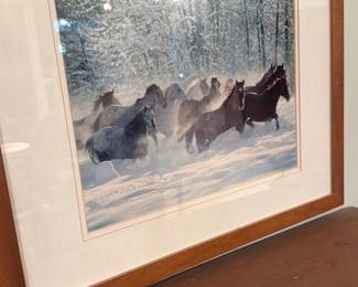 Framed Winter Horses Photograph — Framed color photograph of a herd of horses running through snow with a wooded winter background. The print is double-matted with a gold inner trim and set in a warm wood frame under glass; hanging hardware not shown. Condition appears good with some light surface reflections on the glass and normal wear to the wooden frame edges visible in photos. A decorative, ready-to-hang wall piece that suits living room or entryway display; frame measures roughly typical medium wall-art size.