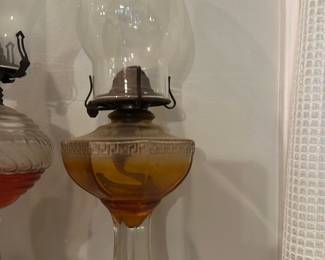 Amber Glass Kerosene Oil Lamp — Tall glass kerosene/coal-oil lamp with an amber-colored font and clear glass pedestal base, fitted with a metal burner and a tall clear glass chimney. The amber font has a decorative band near the top and the clear octagonal pedestal provides stable support; chimney shows no visible chips. Functional style piece for display or light use — burner and chimney appear intact though wick and fuel condition not verified. Overall good condition with minor surface wear consistent with age.