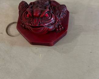 red resin three-legged toad statue — Decorative red three-legged toad/coin frog in molded resin with stylized detailing and inset red glass eyes. The piece sits on an octagonal base and appears to be a decorative Feng Shui-style money toad; underside shows a gold/black maker sticker on stained wood (possible display base). Small surface wear and light scuffs visible on the base and body but overall intact with no obvious breaks. Compact tabletop accent suitable for shelf or mantel display; see photos for exact condition and size.