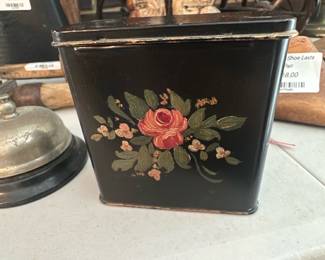 Hand-Painted Floral Tin Canister — Small black metal canister hand-painted with a central rose and leafy floral sprays on the front. The square tin has a fitted lid with light edge wear and a few small surface scratches consistent with age and use; paint remains largely intact. A decorative, functional kitchen or sun-room storage piece that would suit vintage or cottage-style displays. Measures compactly and is easy to tuck on a shelf or countertop.