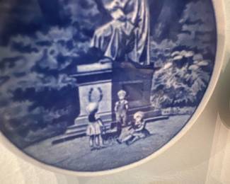 Wall Plate Trio — Set of three blue-and-white Delft-style ceramic wall plates depicting classical scenes: a seated statue with children, a riverside castle, and a mother with child by the shore. Each plate is mounted in a simple white rim and displayed together on the living room wall as shown. Plates appear to be small decorative pieces roughly 6–8 inches in diameter each and light enough for standard wall hanging. In good condition with normal surface glaze wear visible in photos; no obvious chips or cracks noted.