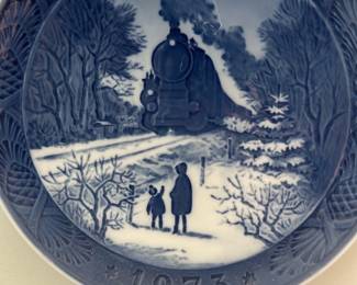 Royal Copenhagen 1973 Christmas Plate — Royal Copenhagen 1973 Christmas collector plate titled "Going Home for Christmas" (Hjem Til Jul) featuring a blue underglaze scene of a steam locomotive with figures in a snowy landscape. The porcelain plate has the factory backstamp, wavy lines mark, painter initials and dated 1973, with a hanging wire attached for wall display. Plate diameter measures approximately 7.5 inches (standard collector plate size), lightweight and easy to display. Excellent collectible condition with no visible chips or cracks in the photos; hanging wire present which may be removed or replaced by the buyer.