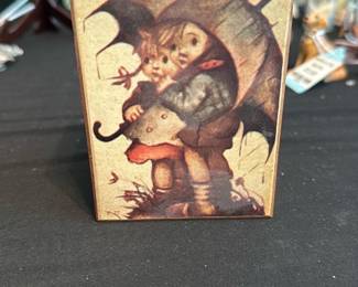 Hummel Music Box “Raindrops Keep Fallin” — Small wooden music/trinket box decorated with a Hummel-style illustration of children under an umbrella on the lid. The interior contains a small wind-up movement (visible) that appears intact but untested; lid hinges are functional and the box shows light surface wear and scuffs consistent with age. Compact tabletop keepsake ideal for music-box collectors or as a decorative jewelry box; see photos for condition and movement detail.