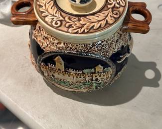 Tilso Lidded Ceramic Jar — Decorative lidded ceramic jar marked "Tilso" on the base, featuring molded handles and a raised scenic medallion with a painted village scene against a speckled ground. The lid has an ornate leaf border and a central floral knob; glaze shows normal surface crazing consistent with age. Good overall condition with no obvious chips or large cracks visible in photos; ideal as a decorative collectible or kitchen accent.
