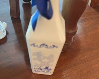 Blue White Ceramic Decanter — Porcelain decanter with blue transfer-style scenic decoration and matching blue ribbon at the neck. Tall rectangular form with a tapered neck and glossy white glaze; decoration shows a traditional landscape motif in cobalt blue. No visible maker's mark in photos; appears intact with light surface wear from display. Measures under 15 inches tall and is a decorative dining-room bottle suitable for display or serving after cleaning.