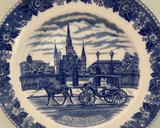 Jackson Square Blue Transferware Plate — Decorative blue and white transferware plate depicting Jackson Square, New Orleans, Louisiana with a horse-drawn carriage scene. The porcelain plate features an ornate floral border and central historic landmark illustration, typical of commemorative tourist ware. Measures approximately 9 inches in diameter (see dimensions) and is suitable for wall display in the living room as shown. In good condition with no visible chips or cracks from the photo; minor surface crazing or light surface wear may be present with age.