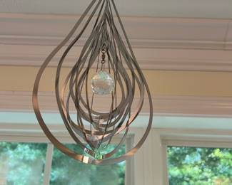 Silver Teardrop Hanging Mobile — Decorative silver-tone teardrop hanging mobile with layered concentric metal hoops and a central faceted glass crystal. Lightweight modern design ideal for a sunroom or window where it catches light and creates subtle movement; shown hanging from a small ceiling hook. Metal shows minimal surface wear consistent with normal use and the crystal is intact with no visible chips. Measures small to medium in size and is ready to hang as-is.