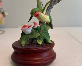 Hummingbird Porcelain Music — Decorative porcelain hummingbird perched over painted flowers on a turned wooden base. Hand-painted polychrome porcelain shows a green body, red throat and detailed wings; the wood base has three small rubber feet and an access hole on the underside (likely for a music box or battery compartment). Good cosmetic condition with no visible chips or cracks in photos and only light surface wear to the base. Compact tabletop accent measuring approximately as shown below.