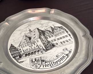 Heilbronn Pewter Commemorative Plate — Decorative pewter wall plate featuring a detailed black-and-white central scene labeled "Heilbronn" with a coat of arms; scalloped rim and an attached hanging tab on the back. The reverse shows a maker's mark (partially legible) impressed into the pewter; plate appears solidly cast with a matte, slightly aged patina. Good wearable decorative condition with light surface wear and small scuffs consistent with age and display; no visible cracks or repairs. Ideal as a European town commemorative or wall-hanging display piece; diameter approximate in dimensions below.