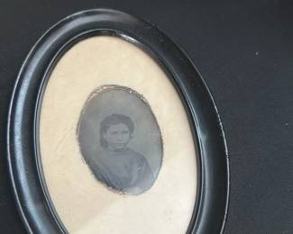 Oval Black Frame Tintype Photo — Small oval black frame containing an early tintype or similar 19th-century style portrait photograph. The faded image shows a seated figure with period clothing and the mat has age toning; frame shows light surface wear and a few scuffs consistent with age. A decorative, display-ready piece for a collection of antique photographs or period decor; exact photographic process and date unknown. Measures small and lightweight—see dimensions below.