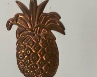 Copper Pineapple Wall Decor — Decorative copper pineapple wall hanging photographed in the sun room. Pressed copper with embossed texture shows a stylized fruit motif and a small top hanging hole for simple mounting. Measures approximately 7 inches tall and 4.5 inches wide, lightweight and easy to relocate on any wall. Good cosmetic condition with light surface wear and natural patina consistent with use; no repairs noted.