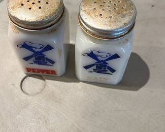 Milk Glass Salt & Pepper Shakers — Pair of milk-glass salt and pepper shakers with blue windmill graphics and red "PEPPER" and faint gold "SALT" lettering. Metal screw tops show surface rust and patina; glass bodies have light staining and expected wear from age. Compact and collectible as a kitchen set or display, both appear structurally sound though tops may need cleaning or replacement. Sold as shown.