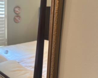 Ornate Gold Wall Mirror — Decorative rectangular wall mirror with an ornate gold-finished carved frame and crest molding at the top. The frame appears to be wood or composite with detailed scrollwork and a warm antiqued gilt finish, perfect for a master bedroom or traditional interior. Overall dimensions approximate a medium-to-large bedroom mirror suitable above a dresser or bed. In good condition with light surface wear to the gilt consistent with normal use; mirror glass looks clear with no visible cracks.