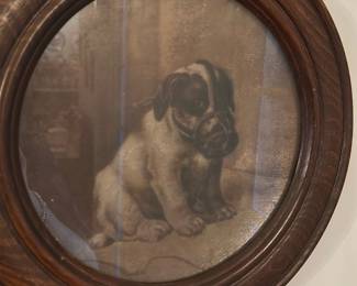 Framed Puppy Portrait Oval Wood Frame — Oval framed puppy portrait in a rounded dark wood frame; the sepia-toned print shows a sitting puppy with visible studio backdrop and glazed front. The solid wood frame has a classic rounded profile with visible grain and light surface wear consistent with age. A decorative wall piece suitable for traditional or cottage-style decor; frame appears structurally sound though glass shows reflections and the print has light age toning. Measures approximately as listed and ready to hang.