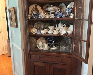 Corner Cherry Wood China Cabinet — Solid cherry corner china cabinet displayed in the living room, featuring a glazed upper display with divided-lite glass doors and a lower storage cabinet with drawer. Warm cherry finish highlights the grain and original wooden knobs; three interior shelves hold china and glassware. Overall sturdy construction ideal for dining or living room display; measures as estimated below. Good vintage condition with minor surface wear and small dings consistent with normal use.