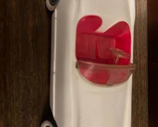 White Toy Convertible Car — Small white plastic convertible toy car with red interior and clear windshield. The model has simple metal axles with rolling wheels and whitewall-style hubcaps; underside shows light wear and one wheel has a blue repair/adhesive. Good playwear with scuffs and minor paint loss consistent with use. Compact, lightweight piece suitable for display or play and easy to store.