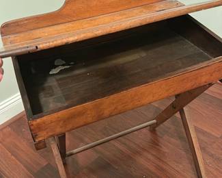 Folding Wooden School Desk — Small folding wooden school desk with lift-top storage and simple X-frame legs. The desk shows original warm patina and surface wear consistent with age, including some scuffs and interior paint residue; the hinge and lift-top function but top sits slightly uneven. Light, compact piece suitable as a decorative accent or small writing table; sturdy but shows age-related wear on legs and finish. Measures approximately 30 in wide by 18 in deep and stands about 29 in tall.