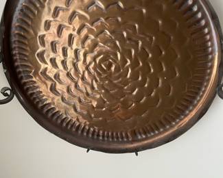 Hammered Copper Wall Tray — Decorative hammered copper wall tray with a radiating petal motif and forged iron hanging loops; photographed in the sun room. The piece features a warm aged copper patina with a concentric embossed pattern and sturdy scroll-style iron handles attached to the rim. Approximately 16 inches across (including handles) and shallow depth suitable as a wall accent or entryway catchall. Shows expected surface wear and small dings consistent with age and use but no major damage; iron hardware has surface rust from age.