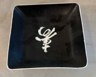 black white porcelain dish — Small square porcelain dish with a glossy black interior and a white abstract character/mark at center. The dish has gently flared sides, a white exterior and a raised circular foot on the underside; photos show light surface wear and minor utensil marks but no chips or cracks. Useful as a sushi soy dish, trinket tray, or decorative accent in the living room. Compact and lightweight for easy display or table use.
